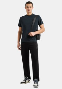 T-shirt nera, maniche corte, con piccolo logo ricamato; abbinata a pantaloni neri; modello che tiene una borsa a tracolla nera; sneakers con dettagli bianchi.