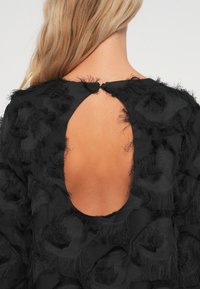 Μαύρο φόρεμα με υφή υφάσματος και φθαρμένες λεπτομέρειες, σταυρωτή πλάτη και κλείσιμο με κουμπί στον λαιμό.