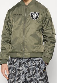 Olive green bomber jacket with a front zip, Raiders logo patch, and five-star embroidery; ribbed cuffs and hem; smooth, shiny fabric.