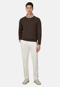 Jeune homme debout, les mains dans les poches, portant un pull marron foncé sur une chemise beige, un pantalon blanc et des baskets beige clair.