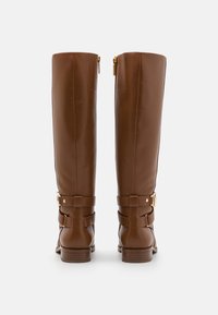 Bottes marron montant jusqu'au genou avec une finition en cuir lisse, détail de fermeture éclair dorée, accents de boucles latérales et talon empilé. Présentent des coutures texturées.