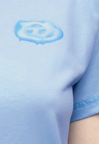 Ανοιχτό μπλε T-shirt με ένα μικρό, αφηρημένο μπλε και λευκό λογότυπο που μοιάζει με σύννεφο στο πάνω μέρος του στήθους κοντά στο μανίκι.