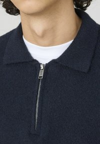 Gilet zippé marine en tissu texturé, avec un col et une fermeture éclair métallique comportant un détail de marque.