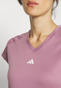 Camiseta deportiva de cuello en V en tela de color rosa claro con mangas cortas. Presenta un pequeño logo blanco en el lado izquierdo. Textura suave, diseño casual.