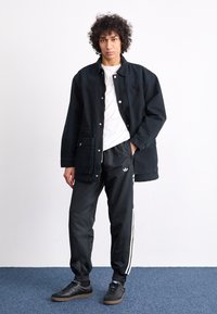 Μαύρο τζάκετ από τζιν με κουμπιά, λευκό μπλουζάκι, μαύρο αθλητικό παντελόνι με λευκές λωρίδες στο πλάι και μαύρα αθλητικά παπούτσια με φόντο καραμέλας.