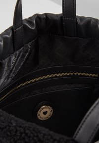 Svart tygväska med en texturerad yta och rundade kanter. Insidan har en slät svart foder med präglade logotyper och en ficka med dragkedja.