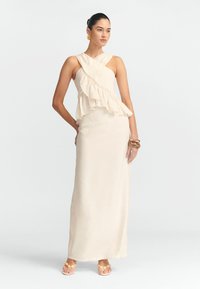 Lang beige kjole med volang, asymmetrisk kant, én skulderstrop og struktureret stof; modellen bærer guld smykker og lyse sandaler.