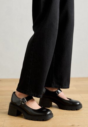 Chaussures Mary Jane noires à talons épais avec des brides à boucle, portées avec un pantalon noir à jambes larges sur un sol en bois.