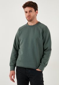 Zelená mikina s kruhovým výstrihom, raglánovými rukávmi a decentným logom na hrudi, vyrobená z mäkkej, textúrovanej látky. Nosená s tmavými džínsami.