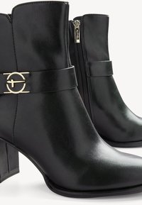 Bottines noires à la finition brillante, avec un accent circulaire doré sur une sangle et une fermeture éclair latérale pour un enfilage facile. Design à talon carré.