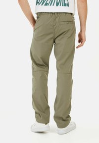 Pantalon olive vert à taille élastique avec une jambe droite, présentant une poche arrière et un tissu lisse et léger. Associé à des baskets blanches.