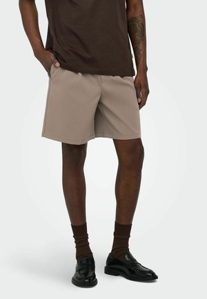 Męski model noszący beżowe krótkie spodenki do kolan, ciemnobrązowe skarpetki, czarne mokasyny i ciemnobrązową koszulkę, stojący z jedną ręką w kieszeni.