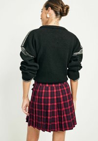 Black knitted sweater with puffed sleeves, accented with silver detailing, paired with a pleated red and black plaid skirt.