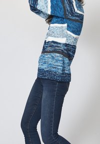 Blå stickad tröja med ett randigt mönster i olika nyanser av blått, vitt och mörkgrått. Löst skuren med ribbad nederkant, tillsammans med mörka jeans.