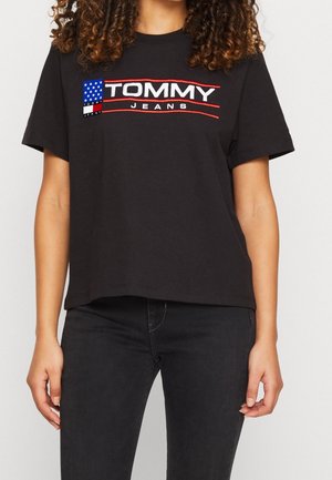 Vrouw draagt een zwart Tommy Jeans T-shirt met logo en een klein Amerikaans vlaggenpatch, gecombineerd met zwarte spijkerbroek.
