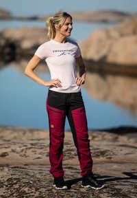 Fitted pink t-shirt with a mountain logo; black and burgundy pants with multiple pockets and contrasting panels; worn with black athletic shoes.
