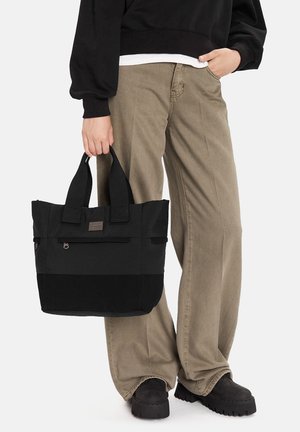 Osoba ubrana w beżowe spodnie i czarne buty trzyma czarną torbę na ramię z dwoma uchwytami, stojąc na tle jednolitego tła.