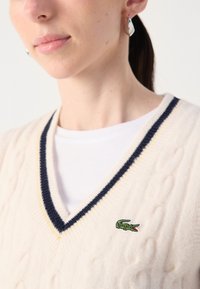 Cremfarbener Kabelstrickpullover mit navy- und gelbfarbigem V-Ausschnitt und einem grünen Krokodil-Logo, das auf der Brust gestickt ist.