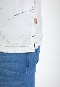 Camicia bianca con texture e motivi marmorizzati neri, dotata di un piccolo etichetta logo e spacchi laterali, abbinata a jeans in denim blu.