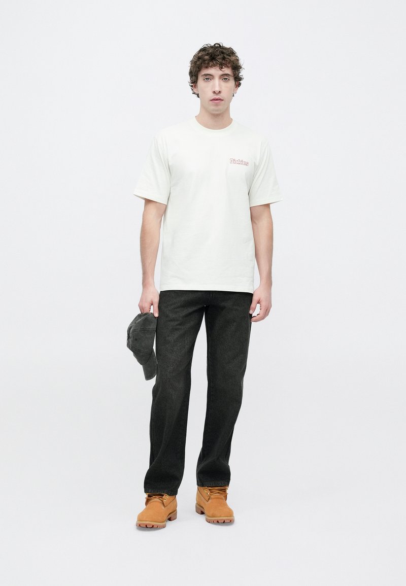 Hvid bomulds-T-shirt med lille rødt logo på brystet, kombineret med mørke jeans, brune støvler og en grå kasket i hånden.