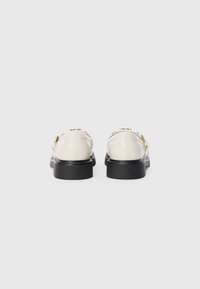 Chaussures en cuir verni beige avec des semelles en caoutchouc noir, dotées d'une bride à la cheville et d'accents en métal doré, vues de l'arrière.