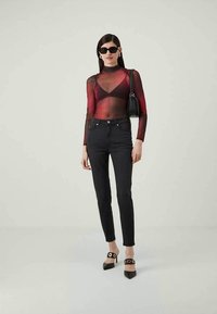 Top largo de manga larga en rojo y negro transparente, jeans negros de cintura alta, tacones negros de punta con acentos circulares y un pequeño bolso negro.