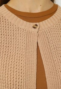 Cárdigan de punto en beige claro con cuello redondo, que presenta un cierre de botón de madera y se lleva sobre una camiseta marrón.