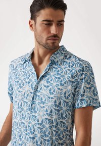 Camisa de manga corta abotonada en tela azul claro con un patrón geométrico blanco, que presenta un cuello extendido y una textura suave.