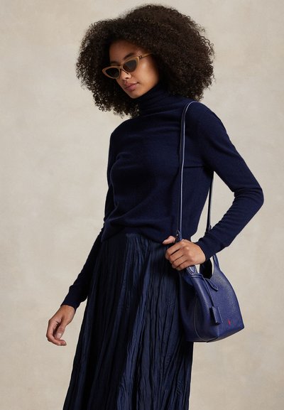 Woman with curly hair wearing brown cat-eye sunglasses, navy turtleneck, pleated skirt, and carrying a small navy handbag on her shoulder.