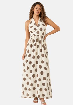 Frau trägt ein ärmelloses, Neckholder-Maxikleid in Creme mit großen braunen Punkten und steht mit einer Hand auf der Hüfte.