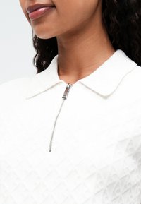 Fehér cipzáros felső textúrált mintázattal, hegyes gallérral és ezüst cipzár részlettel. Az anyag puhanak és könnyűnek tűnik.