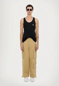 Mees, kes kannab musta ilma varrukat topi, beeži cargo-pükse suurte taskutega, valgeid sneke ja tumedaid prille, seisab tavalise valge taustaga.