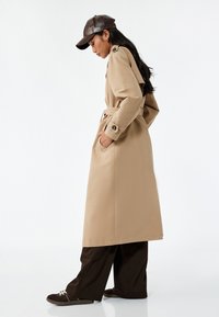 Beiger Trenchcoat mit tailliertem Schnitt, Seitentaschen und Knopfdetails. Kombiniert mit braunen weit geschnittenen Hosen und einer braunen Mütze.