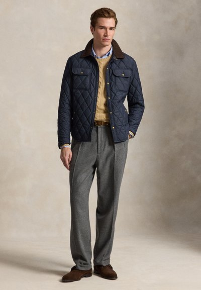 Joven con chaqueta acolchada azul marino, suéter de punto beige con trenzas, camisa azul clara, pantalones grises y zapatos marrones de pie contra un fondo neutro.