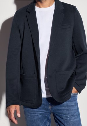 Mand iført en sort blazer over en hvid t-shirt og blå jeans, med den ene hånd i lommen og en ensfarvet baggrund.