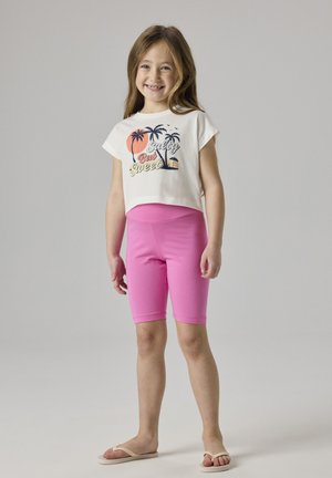 Niña sonriente con camiseta blanca recortada con estampado de palmeras y pantalones cortos rosas hasta la rodilla, de pie descalza sobre un fondo liso.