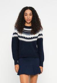 Frau mit lockigem Haar, die einen marineblauen Pullover mit weißen geometrischen Mustern und einen marineblauen Faltenrock trägt, vor einem schlichten weißen Hintergrund stehend.