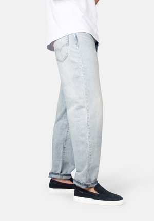 Person trägt hellblaue Jeans und schwarze Slipper mit weißen Sohlen, steht vor einem schlichten weißen Hintergrund.