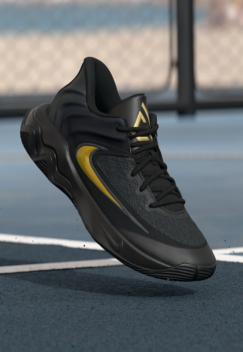 Sorte og guld atletiske sneakers med struktureret stof, snørebånd og tyk sål, der svæver over en udendørs bane med hegn i baggrunden.