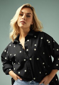 Camicia nera a bottoni con ricamo di margherite bianche, caratterizzata da una vestibilità rilassata e una tasca sul petto. Il design casual si abbina perfettamente ai jeans.