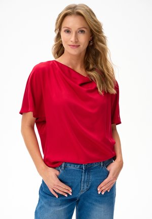 Vrouw met golvend blond haar, draagt gouden oorringen, een losse rode top en een blauwe spijkerbroek, met handen in de voorzakken.