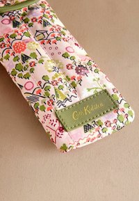 Kvadrična tkanina torbica z cvetličnim vzorcem v roza, zeleni in črni barvi. Vključuje zeleno oznako z zlatim besedilom "Cath Kidston."
