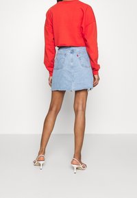 Femme portant un sweatshirt rouge court, une mini-jupe en denim bleu clair avec ourlet brut, et des sandales à talons blancs, se tenant debout devant un fond uni.