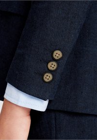 Rękaw granatowego żakietu z trzema ciemnobrązowymi guzikami. Zawinięty mankiet jasnoniebieskiej koszuli widoczny poniżej, ukazujący gładką fakturę i kontrastowy kolor.