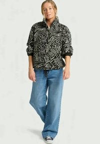 Svart og hvit pull-over fleece med glidelås i kragen, geometriske mønstre og lange ermer, kombinert med lyseblå vide jeans og sorte joggesko.