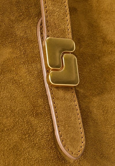 Κοντινό πλάνο από λουράκι μουσταρδί σουέτ δέρματος με ορατές ραφές και χρυσαφί μεταλλική πόρπη σχηματισμένη από δύο αλληλοσυνδεόμενα καμπυλωτά τετράγωνα.