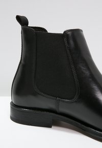 Czarny skórzany botek za kostkę z elastycznymi bokami, panel tekstylny o fakturze oraz zaokrąglonym noskiem. Posiada pętlę do zakładania i gładką podeszwę.