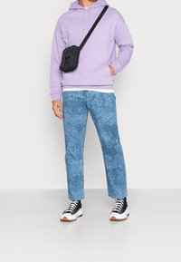 Světle fialová mikina s kapucí, džíny z modrého denimu s texturovaným vzorem, černá crossbody taška a černobílé tenisky s gumovými podrážkami.