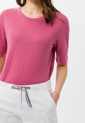 Nainen, joka pukeutunut vaaleanpunaiseen lyhythihaiseen paitaan, joka on laitettu valkoisten housujen sisään. Hänellä on raidallinen vyö, käsi taskussa ja hopeinen rannekoru ranteessa.