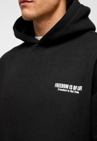 Zwarte hoodie met een zachte textuur, voorzien van witte tekst: "VRIJHEID IS LEVEN" en "Vrijheid Is Niet Gratis" op de voorkant. Standaard pasvorm en ontwerp.